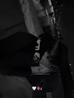 A black and white image showing a couple embracing and kissing, captured from an unconventional angle. The subjects, one wearing a dark hoodie with some visible text, appear in a dimly lit setting. Adjacent to their embrace is a decorative string of lights. An animated heart and teddy bear graphic features in the bottom of the image.