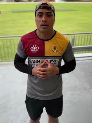 A medium shot shows a man in a multi-colored shirt standing outside. The shirt has a red and yellow block design on the front, with a logo on the red section. The man is wearing a baseball cap and has his hands clasped in front of him. Behind him, there is a green grassy area and metal railings.