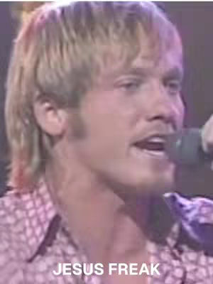 The image is a portrait of a man with fair skin and short, light-brown hair. He is holding a microphone and appears to be singing or speaking passionately. The background is out of focus, suggesting a stage. The words "JESUS FREAK" are written in white text at the bottom of the image.
