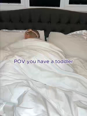 A shot from above, looking down on a bed with white linens. A child with a headband is under the covers. Pink text overlay reads "POV you have a toddler".