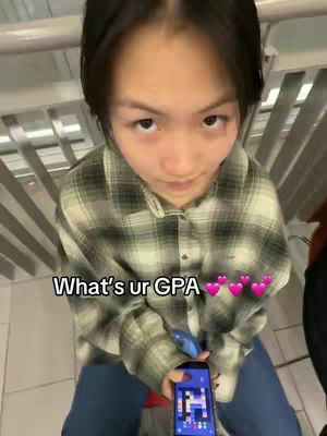 The image shows a person from a high angle, looking directly at the camera. They are wearing a plaid shirt holding a smartphone displaying a game. The image is well-lit, and the setting appears to be indoors, possibly near a staircase or railing.