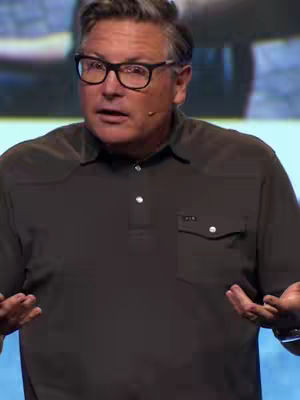 A man in a dark button-down shirt and glasses is captured from the chest up, facing the camera with an open, conversational expression. His hands are outstretched, palms up, as if he's making a point or asking a question. The image is well-lit, with a blurred background. The man has short, graying hair and is wearing glasses with thick black frames.