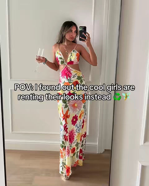 A woman in a floral dress is taking a mirror selfie, holding a champagne flute. The dress is primarily white with a vibrant floral pattern. She is in a well-lit space with white walls and a decorative white door frame. The overlay text indicates the video is about designer fashion rentals and saving money.