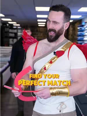 A man dressed as Cupid winks at the camera, holding a bow and arrow. The background is a store interior with racks and shelves.