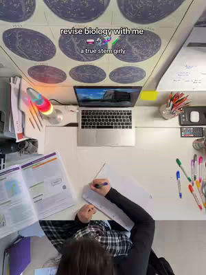 An overhead shot shows a desk setup for studying. The primary focus is on a person writing in a notebook, with studying material like books and stationary around. Above, a world map is visible, and a laptop is open on the desk.