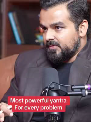 A medium shot of a man with a beard wearing a black suit jacket and a black shirt, sitting in front of a microphone. A text overlay in a red box at the bottom says 'Most powerful yantra for every problem'.