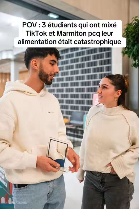 The cover image shows two young adults, a man and a woman, standing side-by-side in a brightly lit indoor space. The man, on the left, is fair-skinned, with short, dark hair and a neatly trimmed beard. He is wearing a light-colored hoodie and holding an object, partially obscured, in his hands. The woman on the right also has light skin, dark hair, pulled back, and is wearing a light colored sweater. They both appear to be looking at each other and smiling slightly. The background includes a textured wall and architectural details. A text overlay at the top of the image provides context.