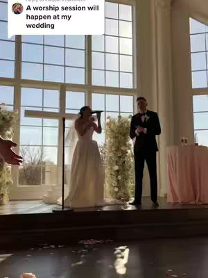 A wedding ceremony taking place in a room with large windows in the background. A woman in a wedding dress is singing into a microphone, while a man in a black suit is standing next to her. The setting looks elegant with floral decorations.