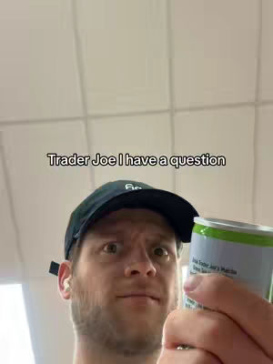 A man with a surprised expression is holding a can of Trader Joe's matcha drink. He's wearing a black cap. The background is a plain, off-white surface.