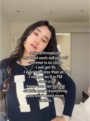A young woman with long, dark hair is centered in the frame, looking slightly up towards the camera. The background is a softly lit room, and she is wearing a dark top with a large white 'H' on it. Text overlays are placed on the image, listing affirmations. The overall composition is well-lit.