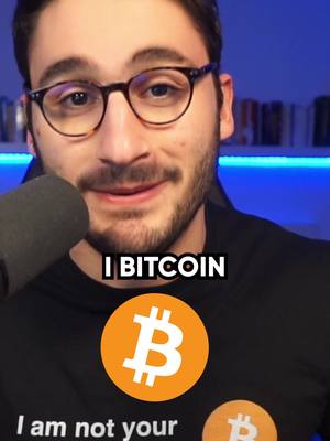 A medium close-up shot of a man with glasses and a beard, with the top of a microphone visible on the left side. Text overlay reads "I BITCOIN" with a Bitcoin logo, and further text at the bottom of a t-shirt.
