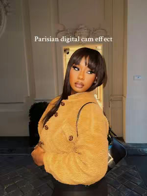 A woman with long black hair and a tan jacket poses in a building with high ceilings and ornate details. The backdrop is of light tan walls and a doorway. The style is modern and aesthetic.