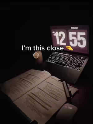 A low-angle shot of a study setup featuring a laptop displaying the time at 12:55, an open book, and a writing utensil. The image is dimly lit.