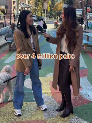 Two women are standing on a colorful, textured surface, possibly a painted street or courtyard. The woman on the left is wearing a casual outfit, while the woman on the right is holding a microphone. The background includes building facades, trees, and other people.