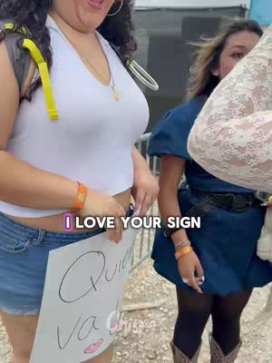 The cover image is a selfie-style shot, capturing two women at an outdoor event. On the left, a woman in a white halter top and denim shorts holds a sign with a custom message, her expression is a mix of surprise and anticipation. To her right, another woman in a blue short sleeve dress and boots is partially visible, holding a cream-colored item and looking towards the sign. A hook text appears on the bottom of the image, with the message "I LOVE YOUR SIGN". The background hints an outdoor setting.