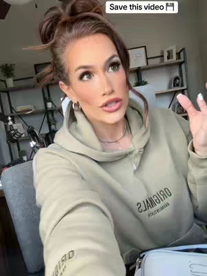 A woman with light skin and brown hair styled in an updo with loose pieces framing her face is the main subject. She wears a light green hoodie. The background features a blurred interior of a room, with glimpses of shelves and décor.