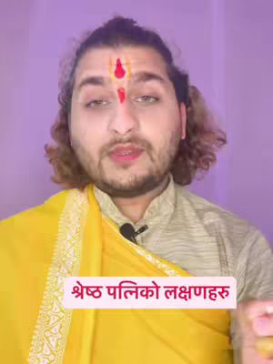 A man with a traditional religious marking on his forehead is centered in the frame, with a light purple background. He seems to be speaking directly to the camera. The man wears a light-colored shirt and a draped yellow cloth. A text overlay in Nepalese language is present.