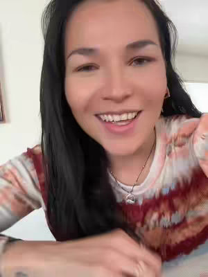 A woman with long black hair smiles directly at the camera against a background of an interior setting. She's wearing a tie-dye patterned shirt in red, white, and pink shades. The composition focuses on her upper body and face, with a necklace visible and some background elements hinted at.