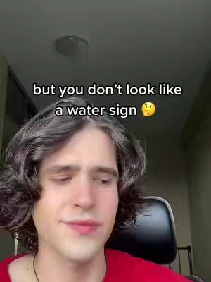 A young man with wavy dark hair is centered in a medium shot. The background is blurred and appears to be a domestic setting, possibly a room with a light fixture visible near the top. He's wearing a red shirt. Text overlayed on the image reads, "but you don't look like a water sign" in white font.