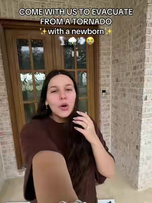 A selfie-style video shows a woman in front of a wooden door. She is wearing a brown sweater and has long, dark hair. The hook text describes the content as they evacuate from a tornado with a newborn. The overall composition suggests a casual, vlog-style approach.