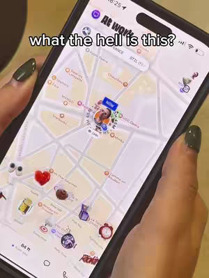 A person holds their phone displaying a map application. The phone is black with a camera in the top center. The app shows a map view with several location markers, including a central one highlighted with a profile picture. Text overlayed on the image above reads, 'what the hell is this?'  The nails are dark green.