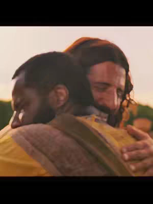 A close-up shot captures two men embracing. The man on the right has long brown hair and clothing that suggests a historical or religious context. The other man has dark skin, and their faces are pressed together in an emotional hug. The background washes out to a bright, warm tone.
