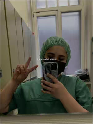 A selfie taken in a medical or healthcare setting, a woman wearing medical scrubs, a green cap, and a face mask is holding up a peace sign with two fingers. White overlay text is present in the image.