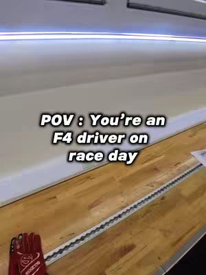 A POV shot with a text overlay. The image is taken from a low angle, looking up at a wooden surface, and a wall. A red racing glove is visible in the bottom left corner. The text overlay reads: "POV: You're an F4 driver on race day". The lighting appears to be fluorescent.