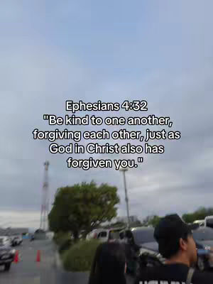 A daytime shot featuring a biblical quote overlayed on an outdoor parking lot scene. The sky is overcast. In the foreground, a person wearing a black cap and a black shirt is visible, with their back to the camera. There are cars and trees in the background.