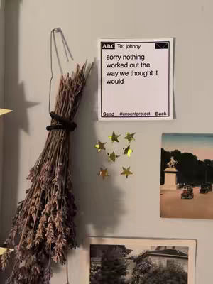 An image showing a wall with various items. A bundle of dried lavender hangs on the left, secured with a black band. To the right, a white card with black text is attached to the wall alongside gold star-shaped decorations. Below, there's a framed picture of a building with trees. The overall aesthetic is neutral and slightly muted.