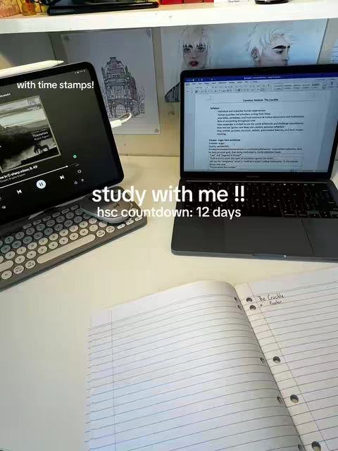 A desk with a laptop and tablet open, alongside some notebooks and a pen. The laptop displays a document, while the tablet shows a music player interface. The setup suggests a study environment with school posters in the background.