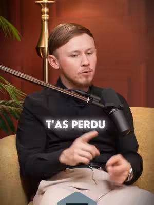 A man with fair skin and short, light brown hair is seated on an off-white padded surface. He wears a black long-sleeved shirt with the words "T'AS PERDU" (You've lost) in white, bold letters across the chest. A microphone extends towards his face. The background includes a reddish-brown wall, a gold-toned lamp, and part of a green plant. His right hand is raised, index finger pointing.