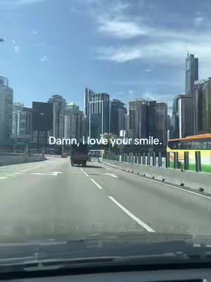 A view from inside a car driving on a highway. The road stretches forward, flanked by buildings on either side, suggesting a city environment. Overhead, a clear, bright sky with scattered clouds fills the background.