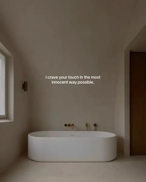 An interior shot of a minimalist bathroom. The room is bathed in soft, warm light and features a large, oval bathtub. The walls and floor have a monochrome color scheme. Text overlayed on the image reads: I crave your touch in the most innocent way possible.