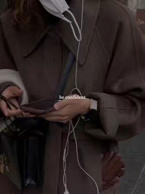 The image features a person, likely a woman, from the chest up, wearing a dark brown coat. The coat has large lapels and a slightly oversized fit. A white face mask hangs from the collar. The person is holding a phone in their hands, with a small purse hanging. The background is blurred, but seems to be an urban setting. The image has a muted color palette, with browns and grayscale tones dominating.