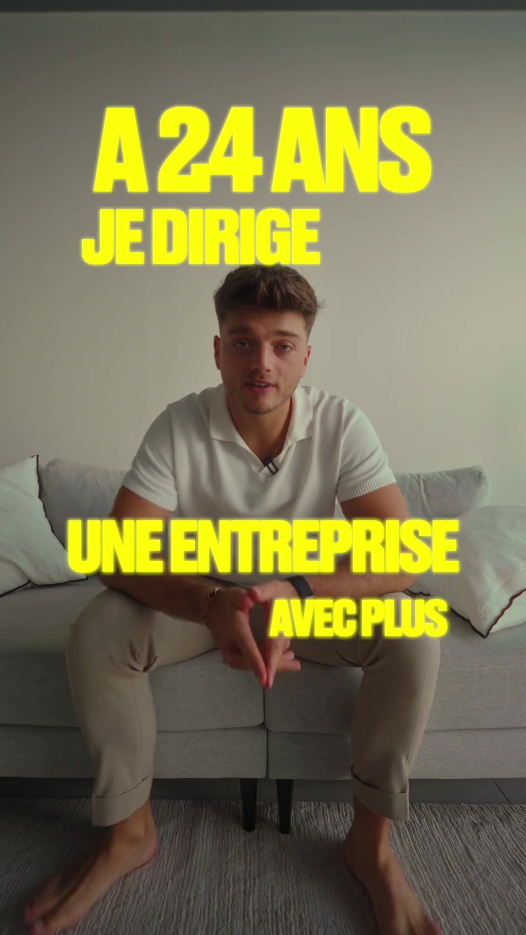 The image features a young man sitting on a couch. Bold yellow text overlays the image, stating "A 24 ANS JE DIRIGE UNE ENTREPRISE AVEC PLUS". He is wearing a white polo shirt and beige pants, the setting has a neutral background and the lighting isn't harsh. The man is looking directly at the camera and is barefoot.