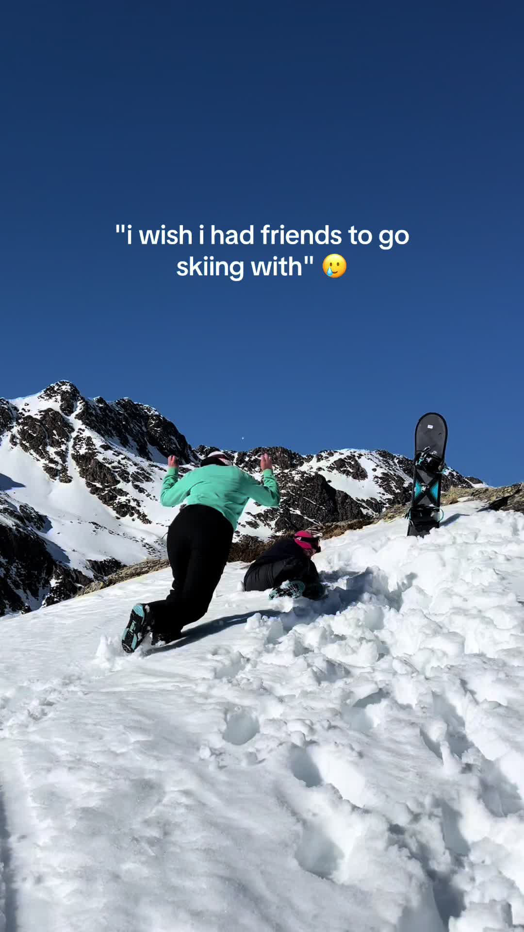 Two people are on a snow-covered mountain, against a backdrop of a clear blue sky and snow-covered mountains. One person is at the top of a snow mound with their arms raised. The person below is sitting on the snow. A black snowboard is in the background.
