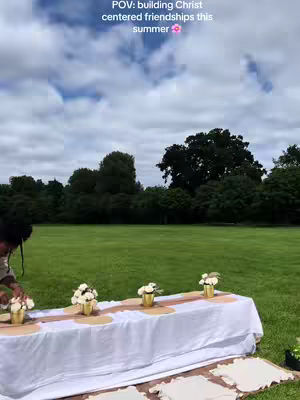 An outdoor shot focusing on a picnic table set for a gathering. A white tablecloth covers the table, with a brown runner down the center. Small gold vases with white flowers are arranged along the table. The scene is set on lush green grass, with a backdrop of trees and a cloudy sky.