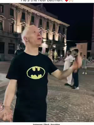 A man in a black Batman t-shirt stands on a paved area at night with a large building in the background, likely in an urban setting. He looks upward with an expression of surprise or anticipation, one arm extended and hand open. Several figures in the distance are partially visible, suggesting a public space.