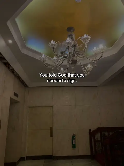 The cover image shows the interior of a building with a high ceiling. A large, ornate chandelier hangs in the center, dominating the frame. The ceiling is a light golden color, possibly with an inset design. The walls are a neutral, beige tone. A door and what appears to be a fire extinguisher are visible along the wall, suggesting an institutional or public space. The overall lighting is soft and indirect, contributing to a somewhat formal or elegant atmosphere. There is text overlay.