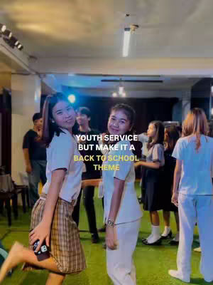A group of young people, likely students or youth group members, are gathered indoors. The image is brightly lit, with the focus on two young women in the foreground. One is holding a phone and is barefoot. The caption overlay announces an upcoming event with a back-to-school theme. The overall impression is one of community, youth, and a welcoming atmosphere.