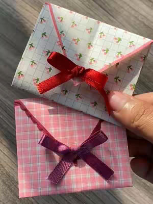 The image showcases two handmade heart-shaped cards or gift boxes arranged on a wooden surface. Each box is crafted from patterned paper and adorned with a ribbon bow. The top box has a white background with a repeating cherry blossom design, and a red ribbon bow. The bottom box has a pink checkered background, with a purple ribbon bow.