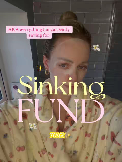 A selfie-style video cover image featuring a woman in a bathroom setting. She's wearing a facial mask and a patterned robe with fruit designs. Text overlays include "AKA everything I'm currently saving for" and "Sinking FUND TOUR". The background is a gray-tiled wall behind the woman.