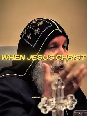 The image features an older man with a white beard, wearing a black hooded garment with ornate embroidery on the hood. Text overlaid in a yellow, glowing font reads 'WHEN JESUS CHRIST'. In the foreground, a glass cross and a water glass are visible.