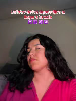 A woman with dark hair and a pink shirt is in the foreground with a text overlay in the background. The text overlay reads "La intro de los signos fijos al llegar a tu vida" followed by zodiac symbols. The woman's eyes are closed, her face is slightly tilted upward. The background appears to be a room with dim lighting.
