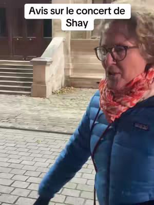 A woman wearing glasses and a blue jacket stands outside, with a building and stairs in the background. Text in a box at the top says, "Avis sur le concert de Shay."