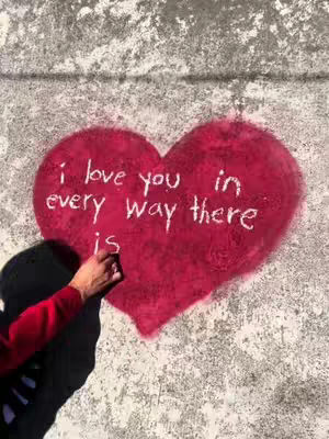 The image shows a red heart painted on a rough, grey wall. Inside the heart, white text reads, 'i love you in every way there is'. A person's hand, draped in a red sleeve, is visible on the left side of the image holding a pen.