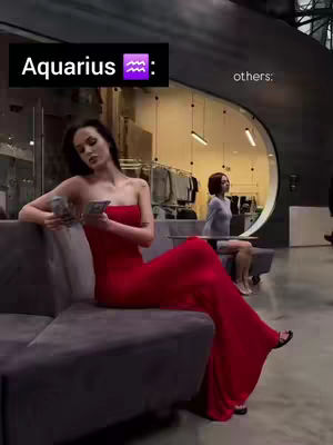 A woman in a red strapless dress sits on a gray couch indoors. She holds cash in her hands and looks thoughtfully to the side. In the background, through a large window, another woman sits on a bench, suggesting a waiting or viewing area. The setting is modern with neutral tones, and the text overlay states "Aquarius".