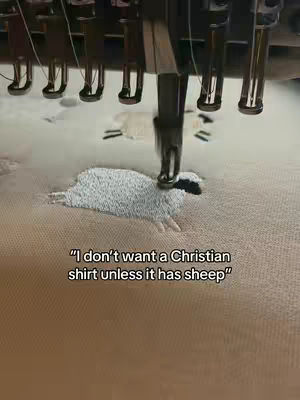An overhead shot shows an embroidery machine in operation. The focus is on a white sheep design being stitched onto a beige fabric. The machine's needle is actively working, and text is overlaid on the fabric.