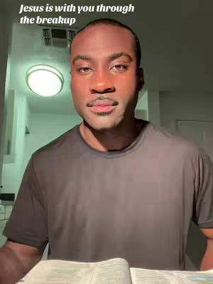 A medium shot of a Black man with a neutral expression. The lighting is soft, and he appears to be indoors, possibly in a room with a light fixture on the ceiling. In the background, there's a glimpse of a hallway or another room. The light is positioned to illuminate his face. He is holding a book open, which suggests a focus on religious content, given the text overlay.
